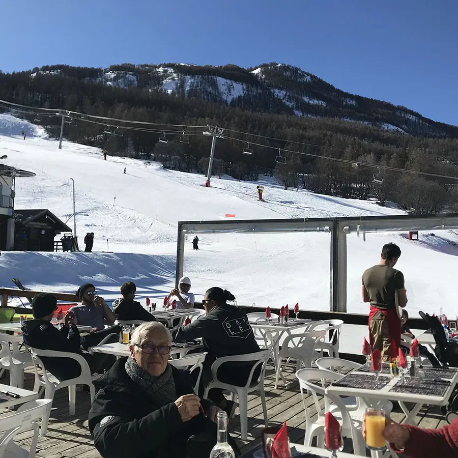 Terrasse extérieure du restaurant en hiver, tables et chaises en plastique, vue sur les pistes enneigées et les montagnes, télésiège à proximité