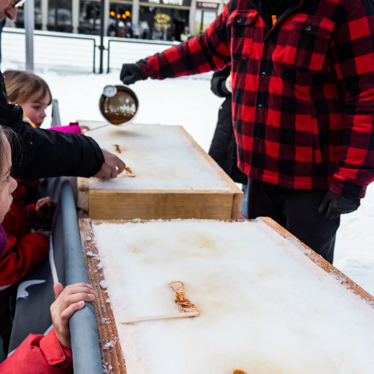 On voit des enfants s'initier au vélo-ski sur la neige avec l'aide d'adultes. À côté, une animation gourmande permet de déguster de la tire sur neige en enroulant du sirop chaud sur des bâtonnets.