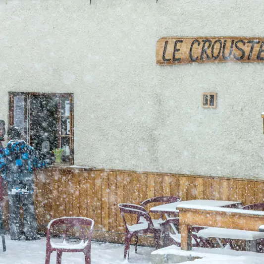 Detail of the restaurant's facade with the wooden sign Le Croustet under the snow, tables and chairs