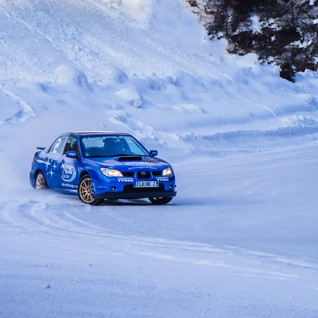 Cette image montre une voiture de rallye bleue en train de déraper sur un circuit de glace à Flaine. La voiture soulève une fine poudre de neige en négociant un virage.