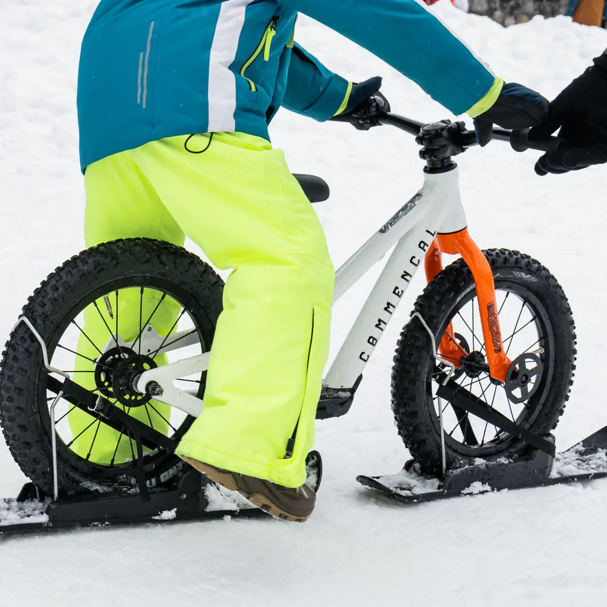 Un enfant portant un pantalon jaune fluo et une veste bleue est assis sur un vélo-ski blanc. On voit bien les patins noirs fixés sous les roues pour glisser sur la neige.