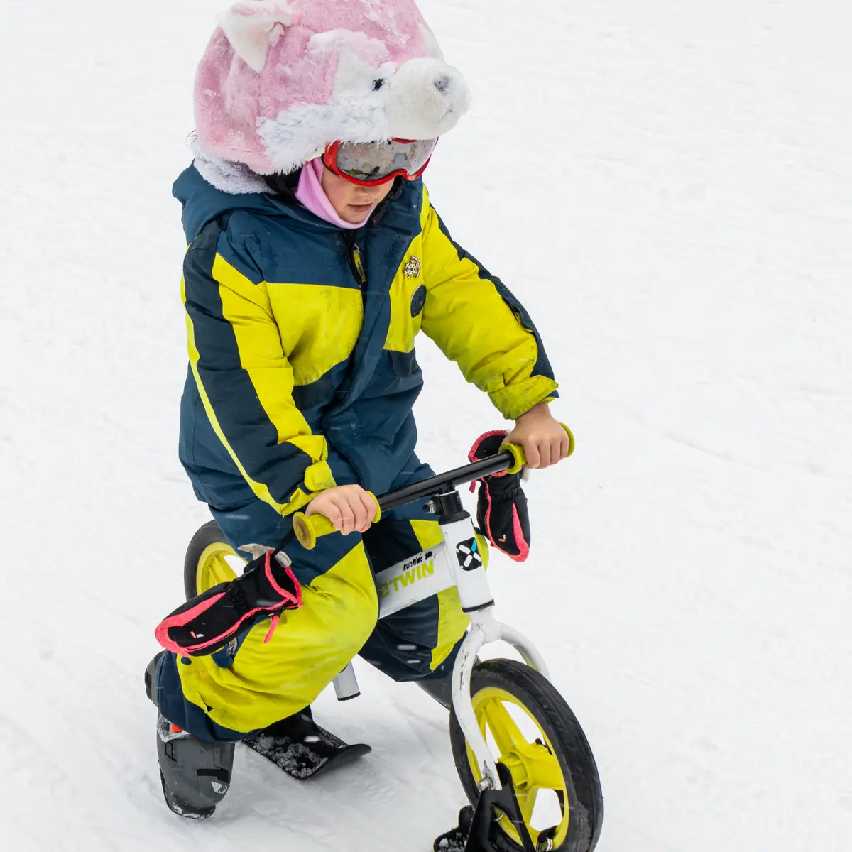 Un jeune enfant portant une combinaison de ski colorée et un bonnet rose en forme de tête d'animal descend une pente de neige sur un petit vélo-ski blanc.