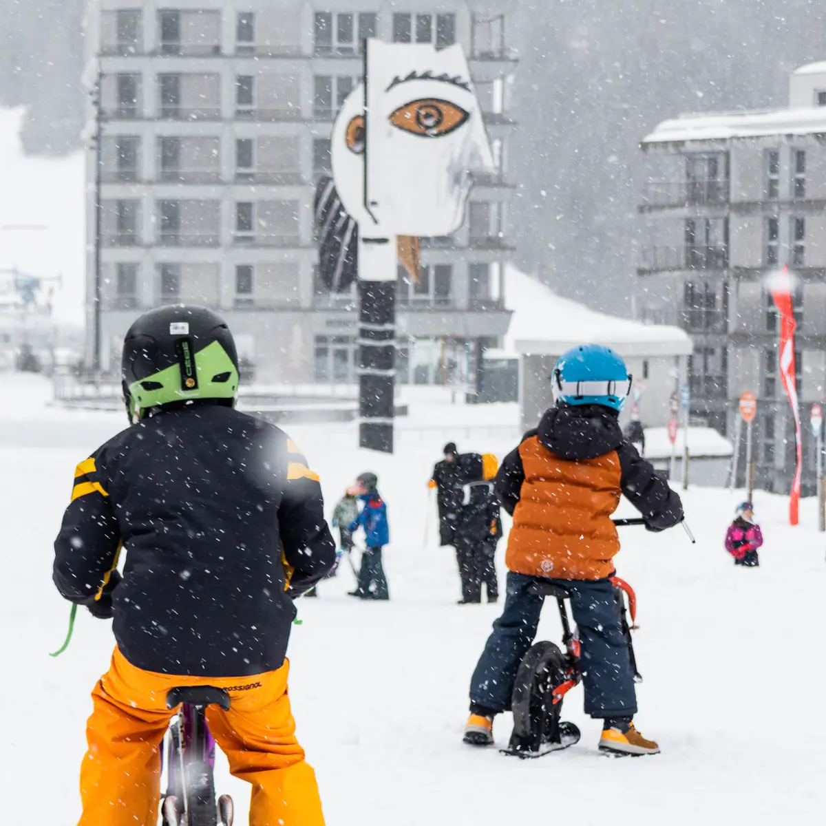 Deux enfants de dos, équipés de casques et de vêtements de ski colorés, font du vélo-ski sur une piste enneigée.