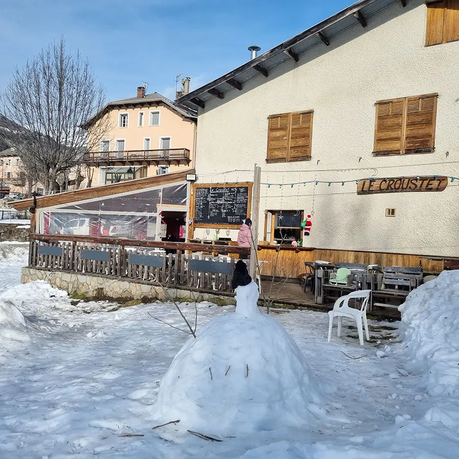 Overview of the restaurant in winter: a two-story stone and wood building with a wooden terrace on the ground floor, tables, and chairs. Snow-covered meadow in front of the restaurant. Wooden sign reading Le Croustet.