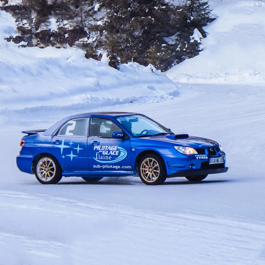 Cette photographie capturent une berline de sport bleue lancée à vive allure sur le circuit de glace de Flaine, entouré de sapins et de parois rocheuses enneigées.