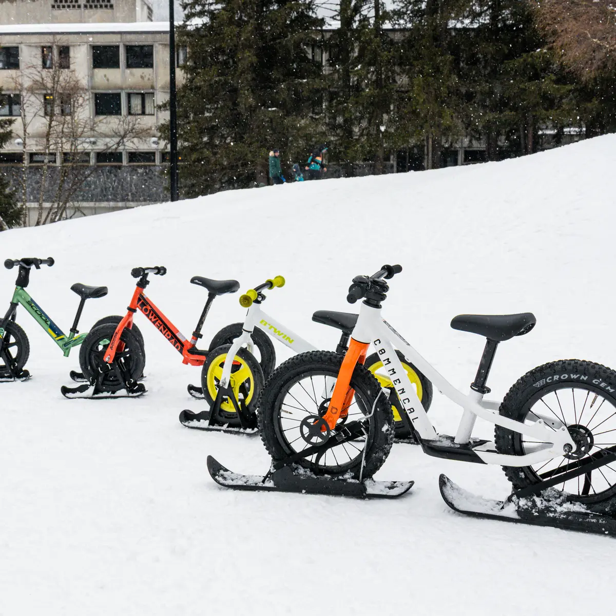 Quatre vélos-ski de différentes couleurs (vert, rouge, blanc et jaune) sont alignés dans la neige sur une pente douce.