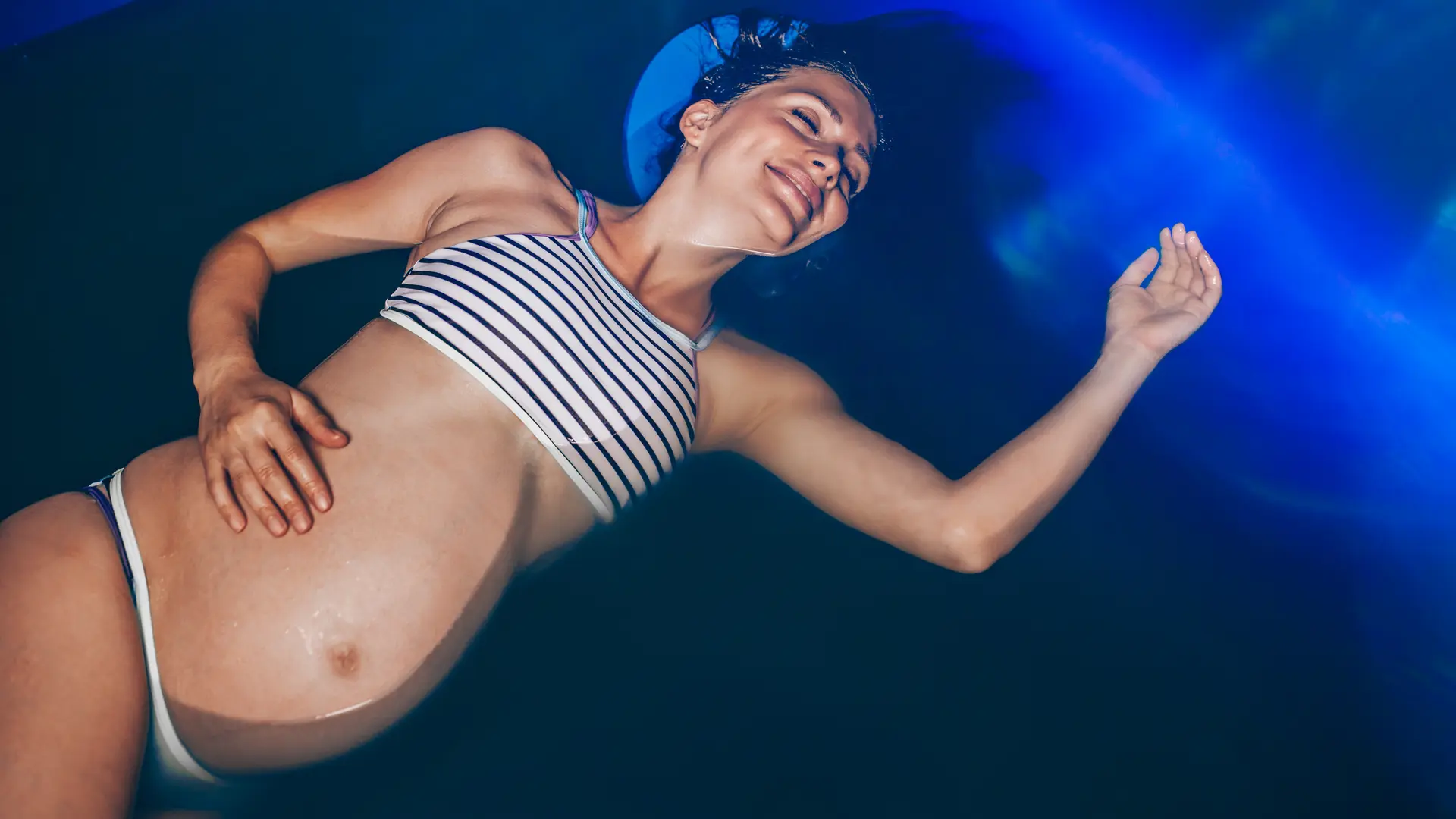 Beautiful pregnant woman floating in tank filled with dense salt water used in meditation, therapy, and alternative medicine.