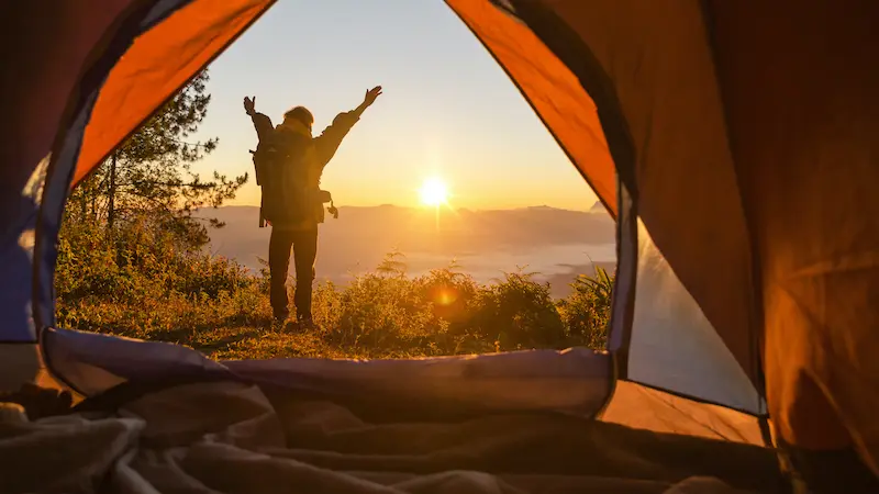 Hiker stand at the camping front orange tent and backpack in the mountains. Backpacker looking into the distance