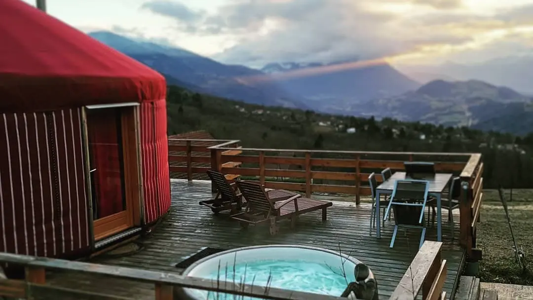 Une yourte rouge avec une vue dégagée sur les montagnes. Sur la terrasse, deux chaises longues en bois et une table pour 4 personnes.