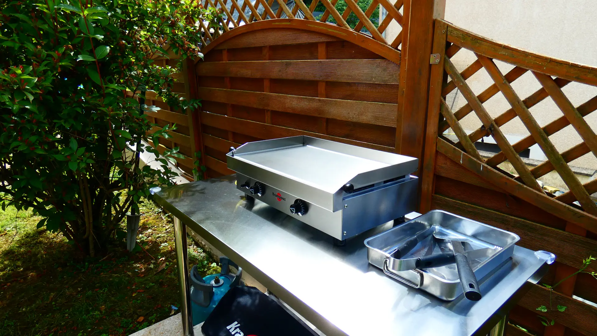 En éxtérieur, sur une table en inox, un appareil à plancha, sa bouteille de gaz et un plateau avec une spatule et une paire de pinces se trouvent à côté d'un arbuste.