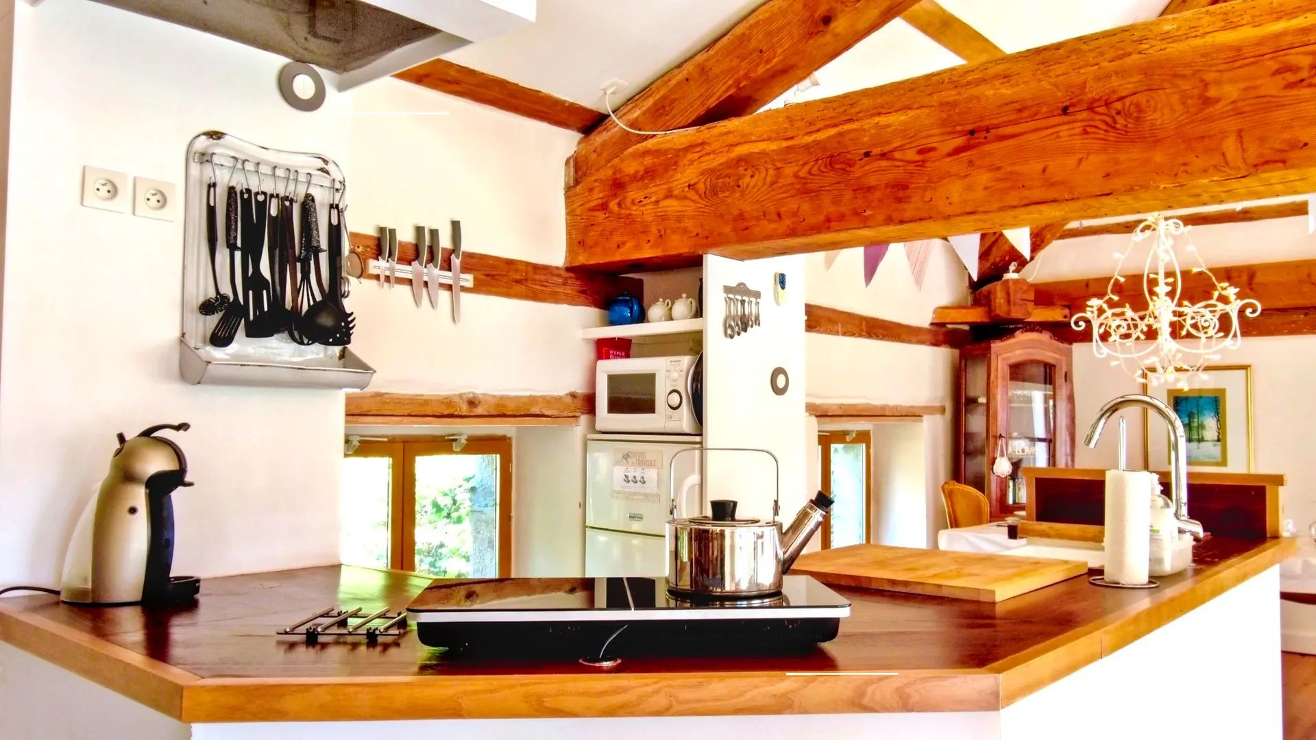 La cuisine se trouve entre la salle à manger et le salon de la mezzanine. Une petite cuisine américaine avec le nécessaire pour cuisiner. Le bar en bois est de forme octogonale ouvert avec un évier, une table à induction et sa bouilloire et contre le mur, une machine à café. On aperçoit des ustensiles de cuisine accroché à un support ancien détourné. A droite, une niche avec une étagère comprenant trois théières. En dessous, il y a un micro-onde et une réfrigérateur (frigo et une partie congélation). Au dessus de la fenêtre, on trouve un support magnétique pour ranger les couteaux de cuisine. La luminosité de la mezzanine est apportée par la lumière traversante venant des fenêtres.