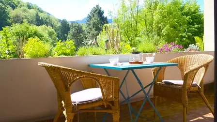 La terrasse est meublée d'une table en métal et de deux chaises en osier avec un arrière-plan boisé et vert.