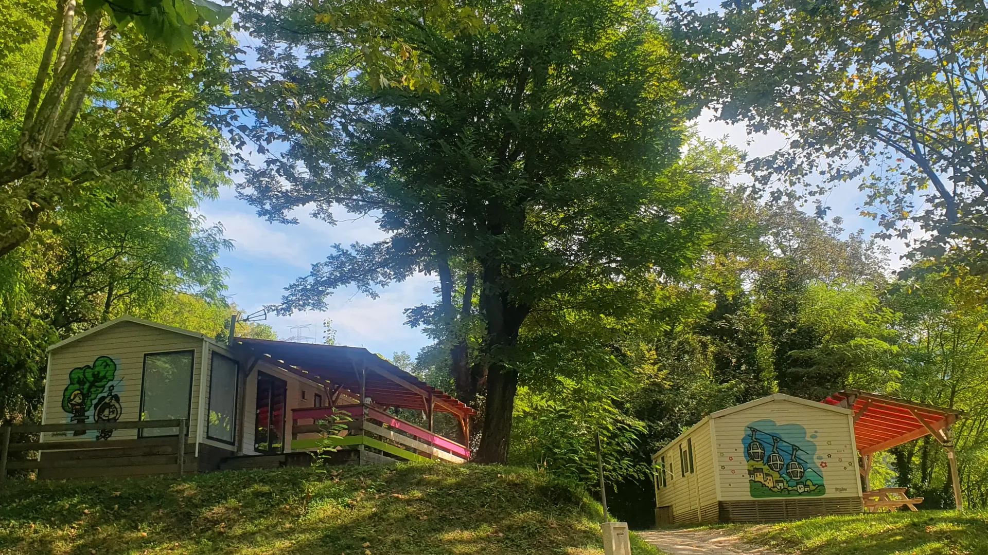 On peut voir un camping avec des hébergements de loisirs en bois, situés dans un environnement boisé. Un chemin de terre serpente entre les différents emplacements.