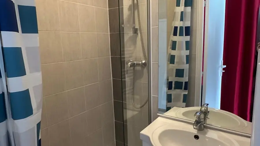 Il s'agit d'une salle de bain carrelée avec une douche partiellement fermée par une paroi de verre et un rideau à motifs à carreaux bleus, blancs et turquoise, faisant face à un meuble-lavabo blanc surmonté d'un miroir.