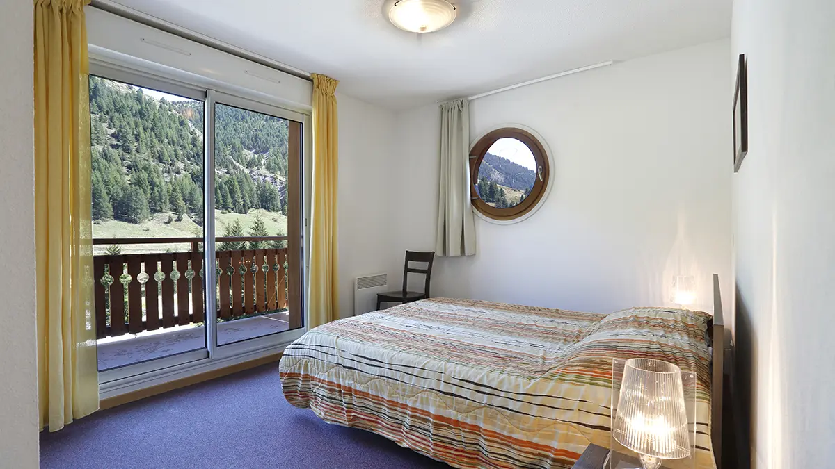 Chambre à coucher d'appartement avec lit double, fenêtre hublot, porte-fenêtre donnant sur balcon