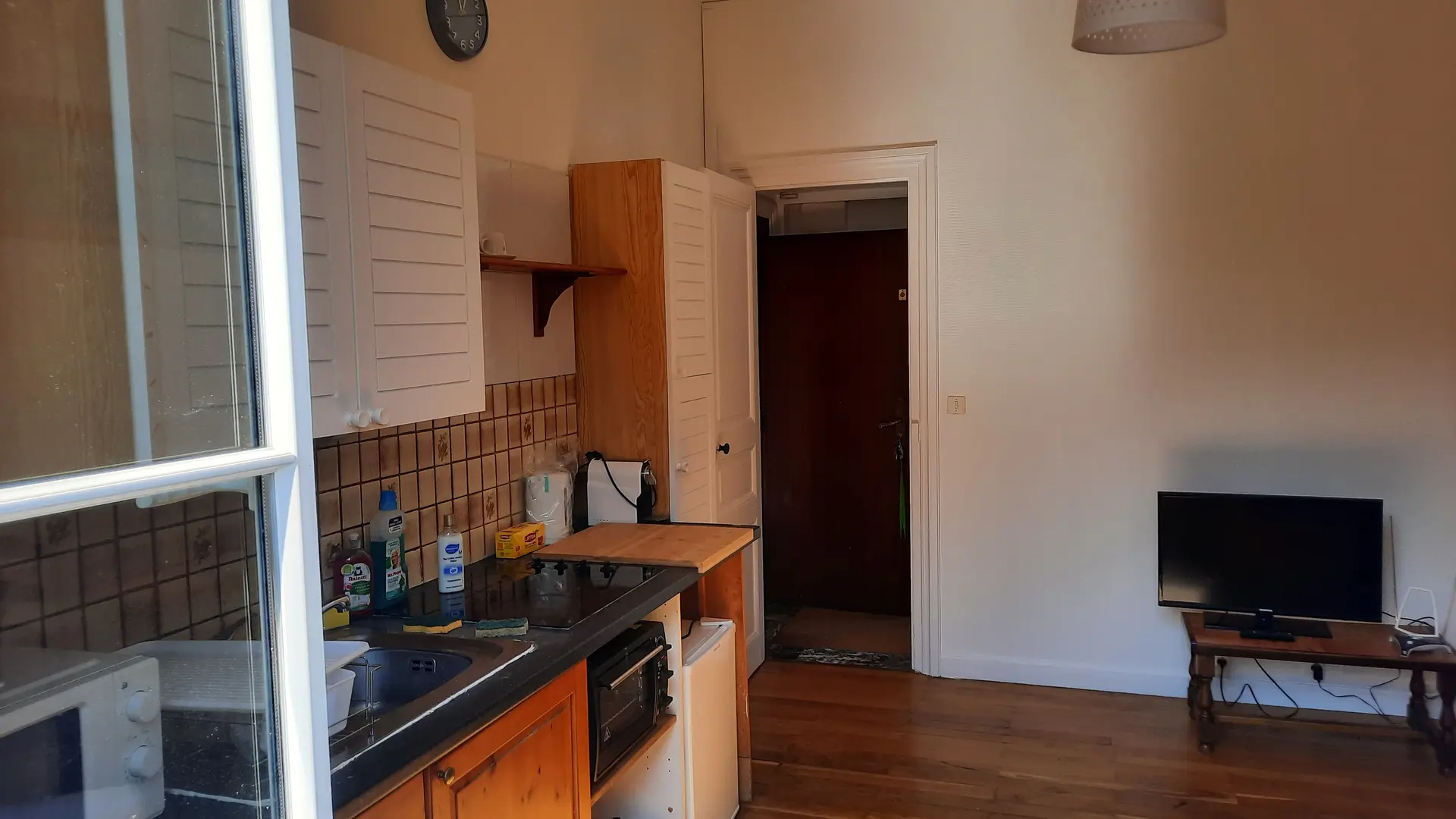 Cette pièce possède un sol en parquet, comprenant un coin cuisine sur la gauche et un espace de vie avec une télévision sur la droite, avec une porte ouverte au fond menant à la chambre.