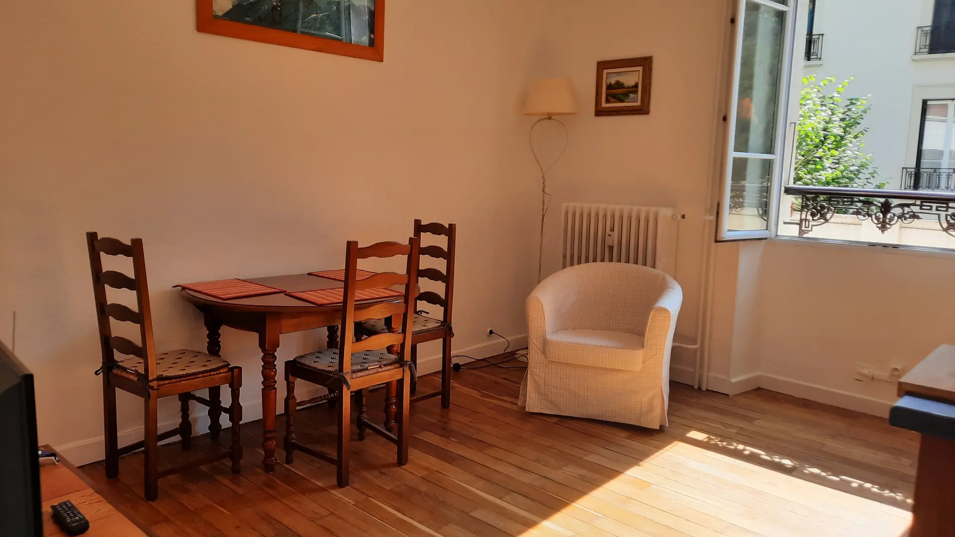 Cette pièce de vie, dotée d'un parquet au sol, est meublée d'une table et de chaises en bois sur la gauche, ainsi que d'un fauteuil placé dans le coin droit à côté d'une grande fenêtre ouverte.