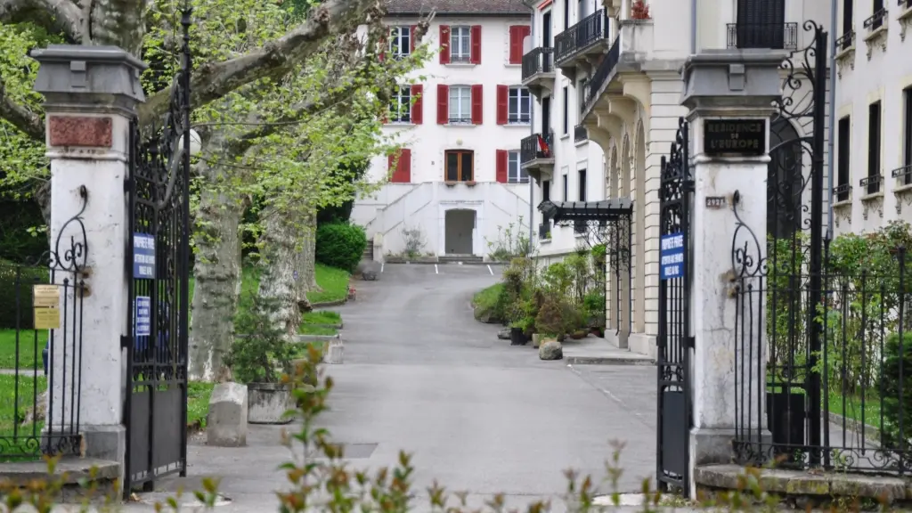 L’image montre l’entrée d’une résidence nommée Résidence de l’Europe, avec un portail en fer forgé ouvert donnant sur une allée bordée d’arbres et menant à un bâtiment blanc aux volets rouges.