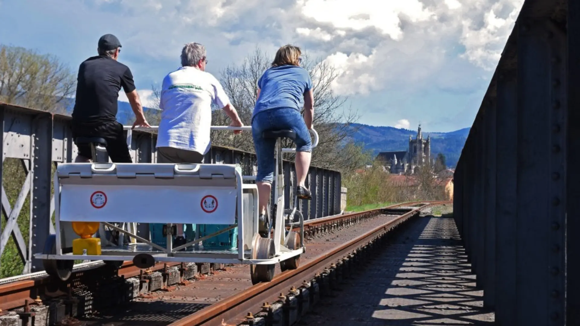 Trois personnes sont sur le vélorail d'Agrivap et traversent un pont. Ils reviennent à Ambert car on aperçoit sur la gauche l'église d'Ambert ainsi qu'une partie des montagnes avoisinantes. Il fait beau avec un ciel ennuagé.