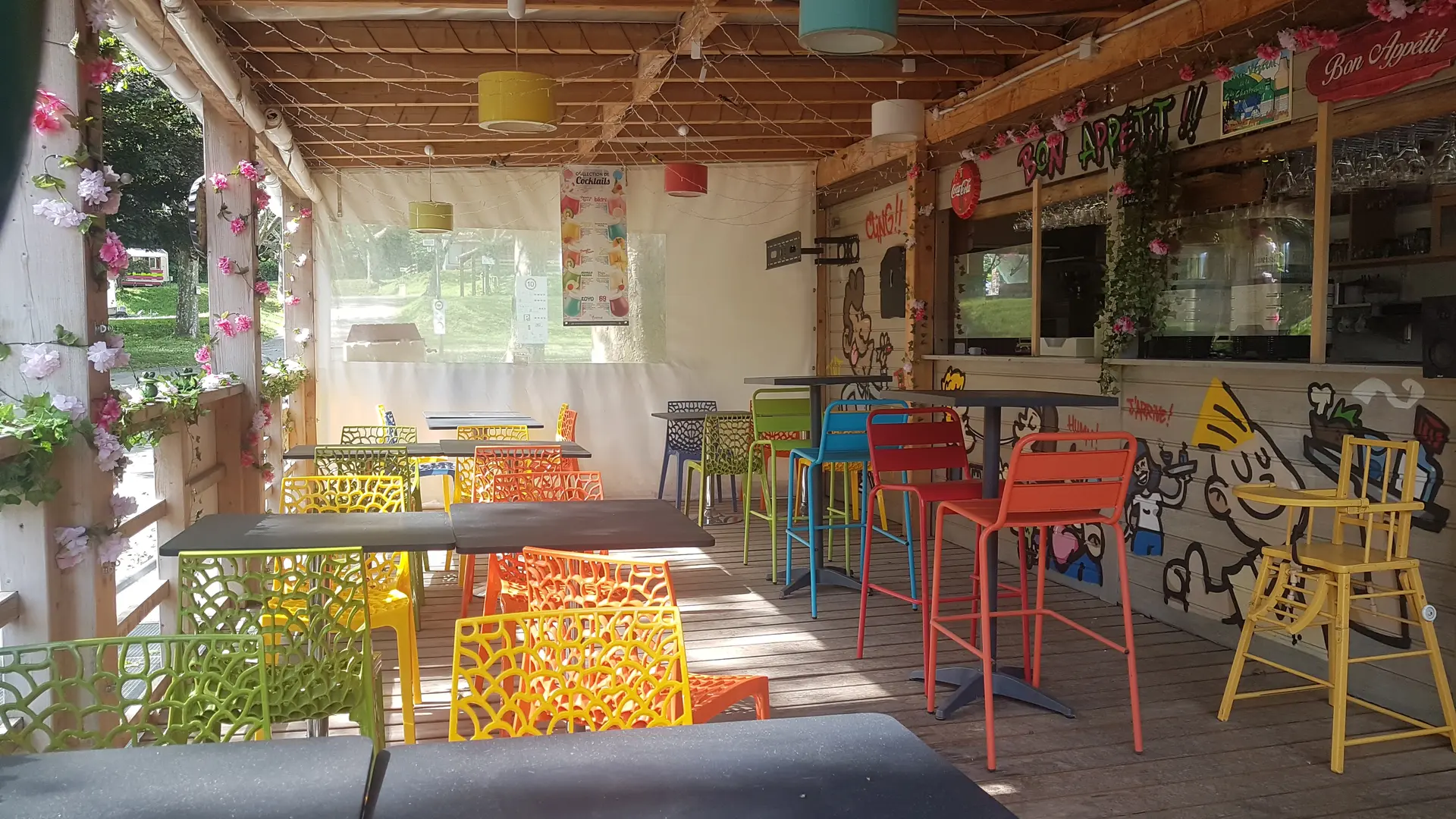 espace de restauration couvert en bois, comprenant un comptoir et des tables avec chaises colorées, situé dans un environnement extérieur.