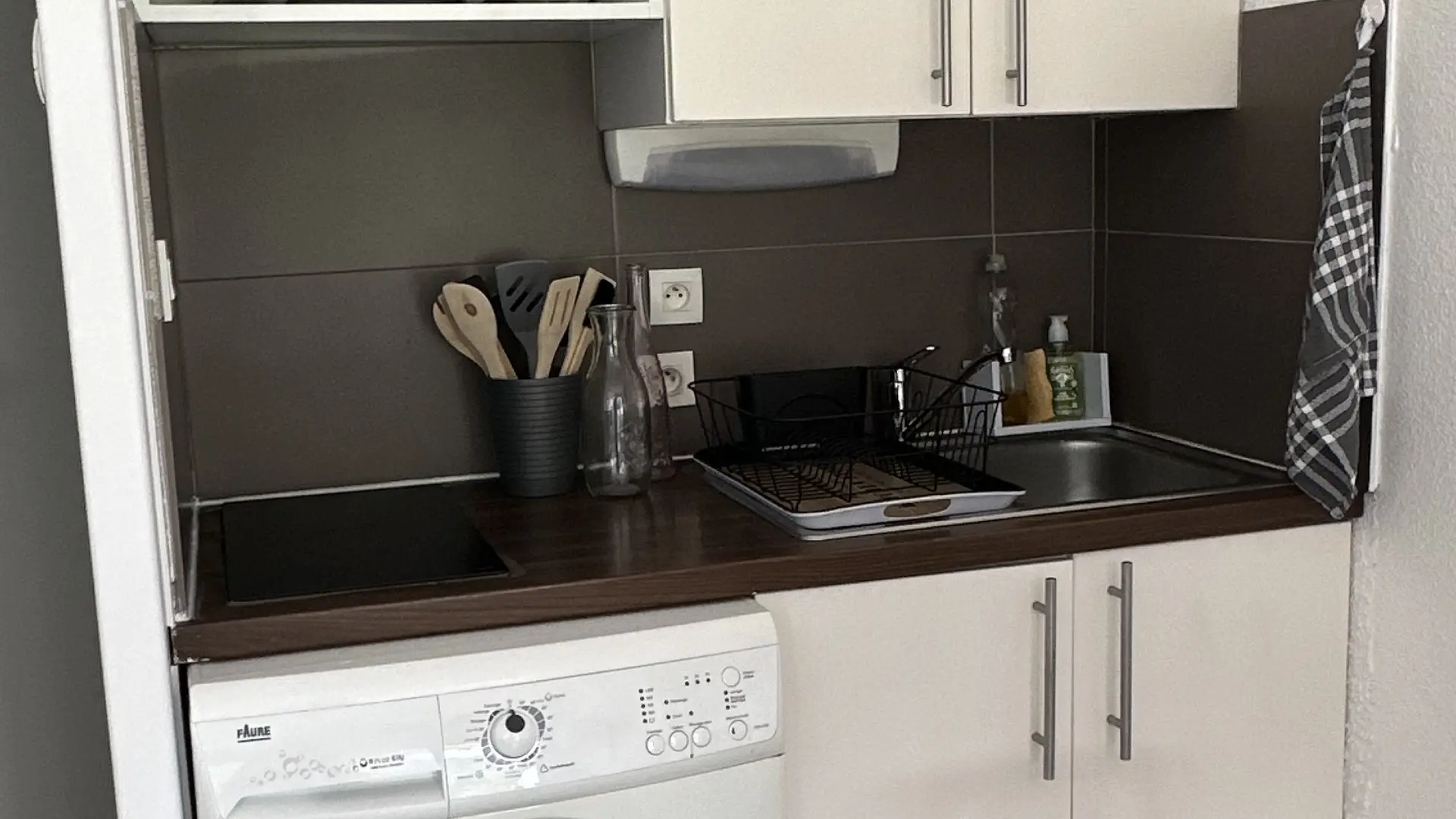 Un coin cuisine équipé d'une machine à laver blanche sous un plan de travail foncé avec un évier et des plaques vitrocéramique, et surmonté de meubles hauts clairs, avec un micro-ondes encastré dans un meuble.