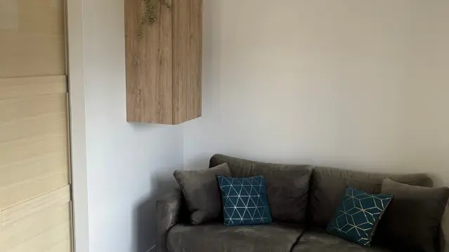 Un canapé gris foncé avec des coussins bleus est disposé dans un coin du salon, accompagné d'une petite table basse carrée blanche posée sur un sol clair.