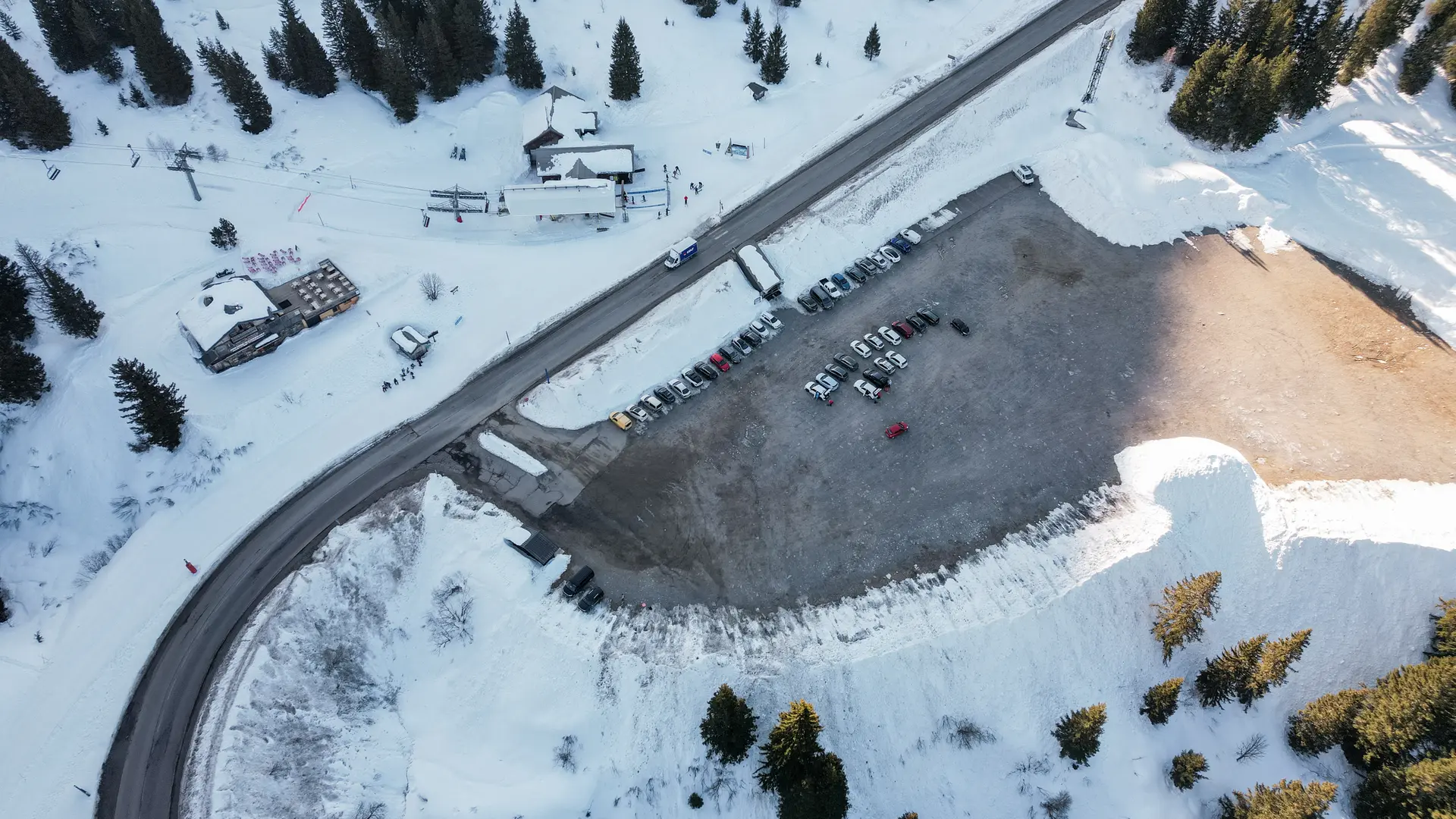 Een skigebied vanuit de lucht met een parkeerplaats in de sneeuw. Een weg loopt langs enkele chalets en skiliften.
