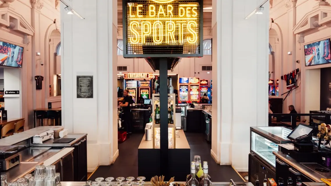 Le bar des Sports du casino JOA d’Uriage est entouré de hauts tabourets confortables. Derrière des arches blanches, on aperçois les machines à sous au fond.