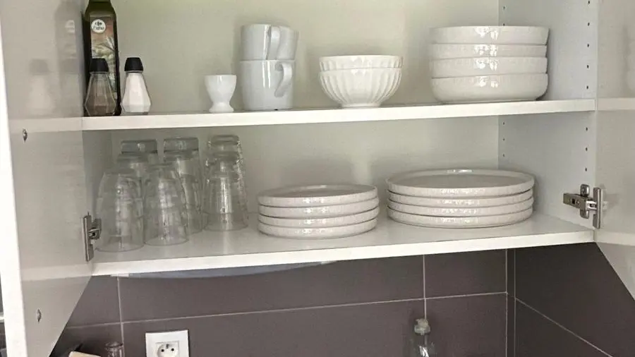 Cette image présente une étagère ouverte de cuisine remplie de vaisselle blanche et de quelques autres objets, positionnée au-dessus d'un plan de travail foncé où sont posées des bouteilles et d'autres ustensiles.