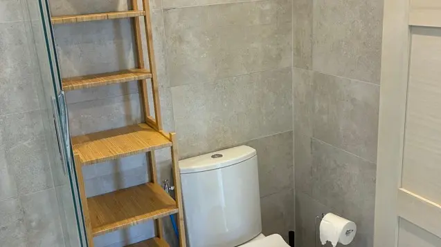 Des toilettes blanches sont installées à côté d'une étagère étroite en bois clair, le tout dans un espace aux murs et au sol carrelés de gris.