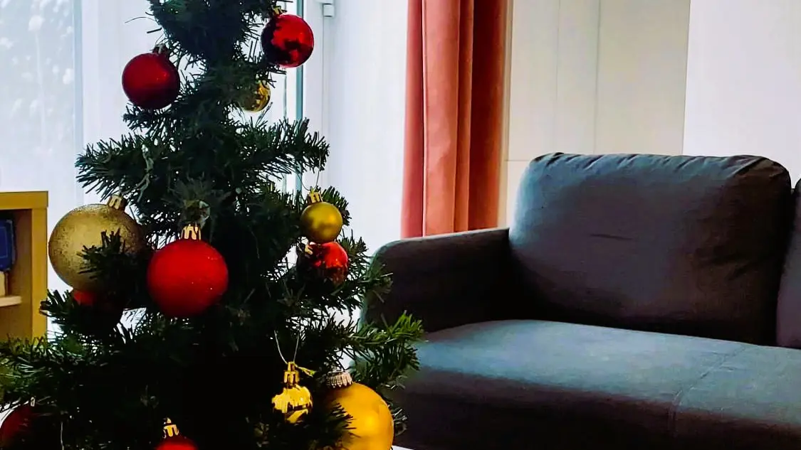 Un petit sapin de Noël décoré posé sur une table basse en premier plan, avec un canapé gris sombre, des rideaux rouges et une grande fenêtre en arrière-plan dans un salon.