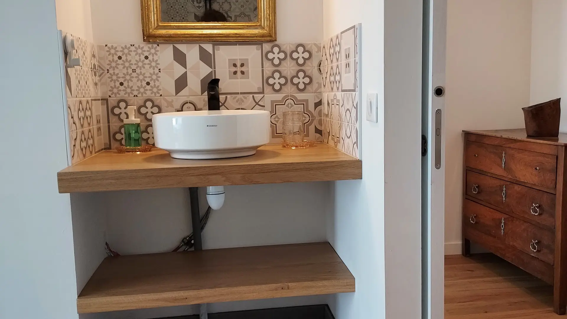 Au premier plan, on peut voir un lavabo en porcelaine blanche posé sur un meuble en bois. Le meuble est doté de deux étagères ouvertes, également en bois. Derrière le lavabo, un miroir rond est accroché au mur.