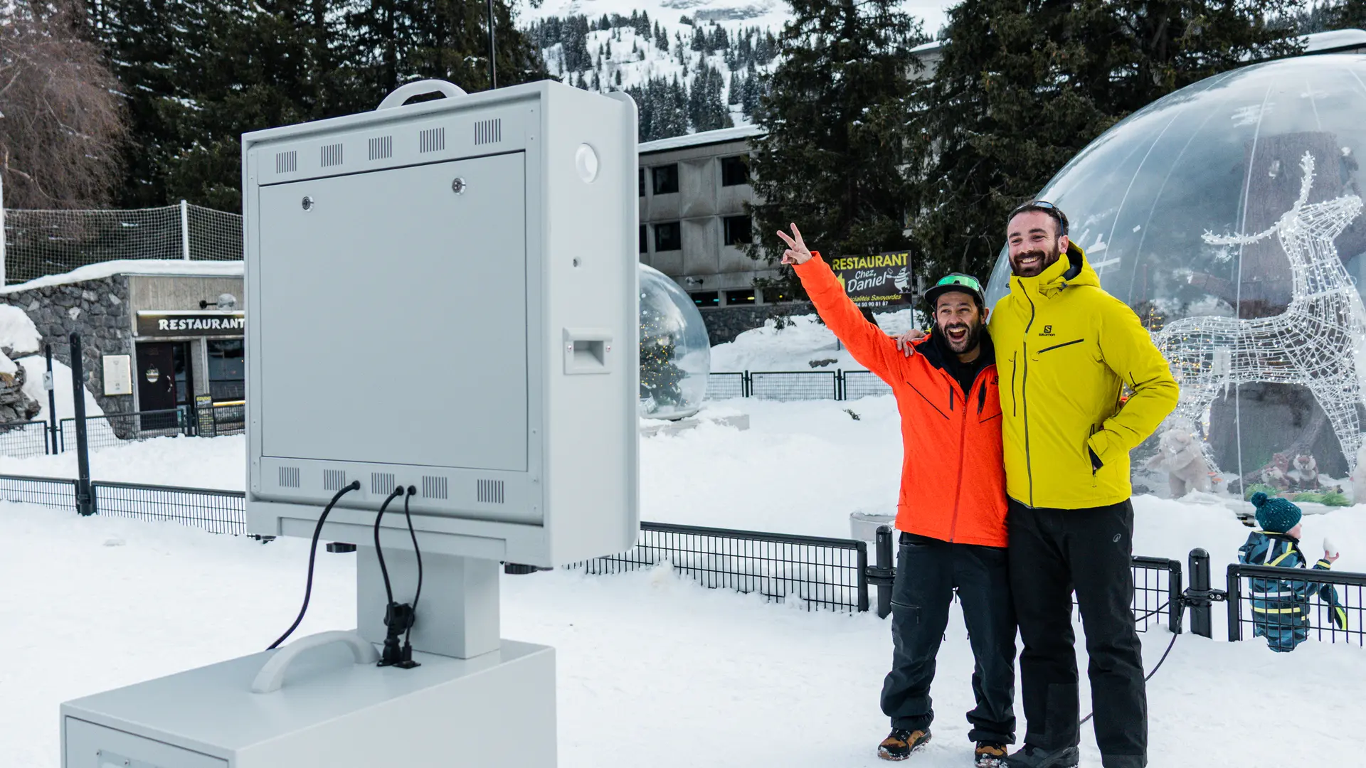 Deux hommes en tenue de ski sourient et posent devant une borne photo installée dans une station de montagne enneigée.