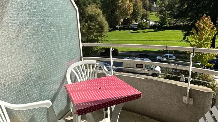 Un balcon meublé d'une petite table carrée recouverte d'une nappe rouge à pois blancs et entourée de deux chaises de jardin blanches en plastique, le tout donnant su le parc Thermal d’Uriage en contrebas.
