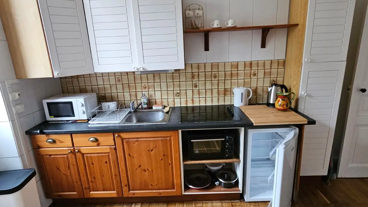 La pièce est une cuisine avec des meubles bas en bois clair et des placards muraux blancs, surmontée d'un plancher en bois et d'un mur de couleur claire.