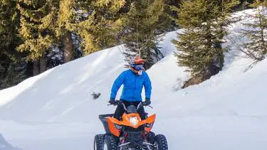 Une personne portant une veste bleue et un casque orange conduit un quad orange et noir sur un chemin enneigé.