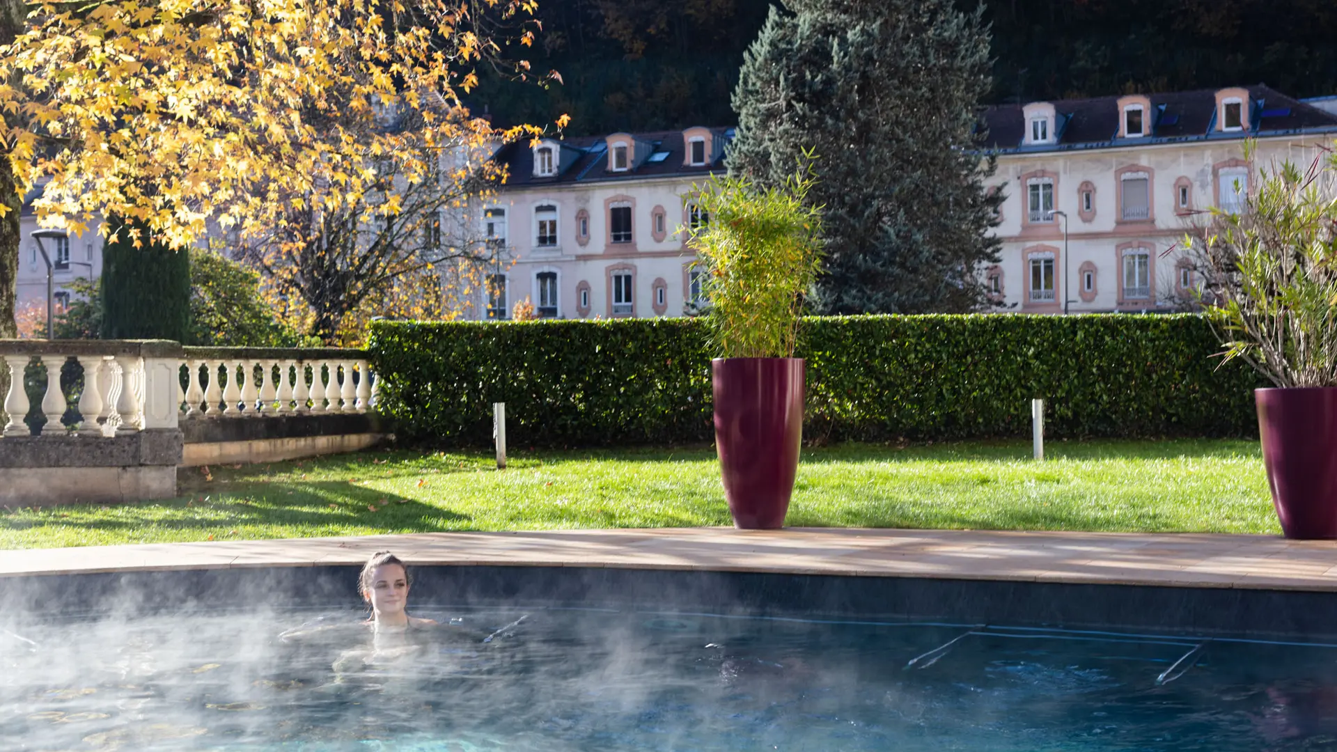 L'image présente un espace aquatique extérieur avec une piscine au premier plan. À l'arrière-plan, on distingue une haie de buissons touffus et, au-dessus, on aperçoit les bâtiments de l’avenue des thermes d’Uriage.