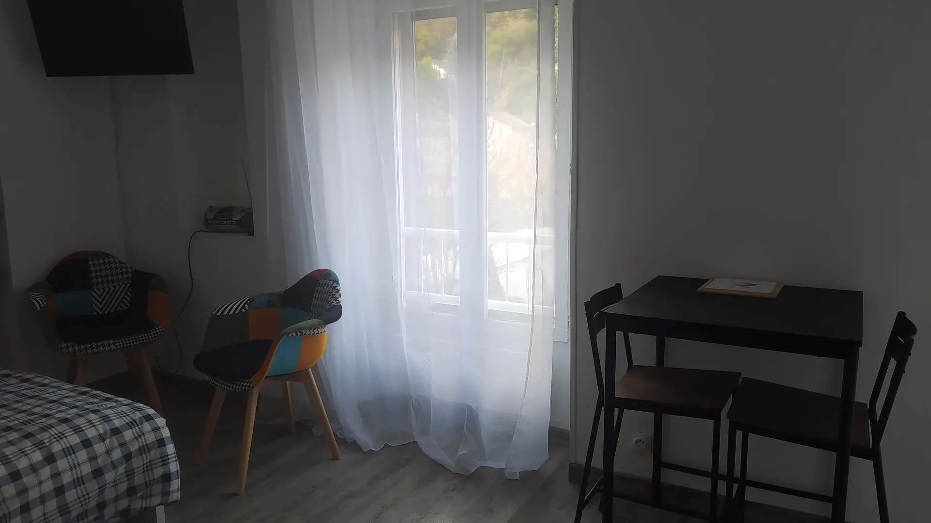 Un studio meublé de façon simple et moderne, avec un lit, une table et des chaises, éclairé par une grande fenêtre à rideaux blancs.