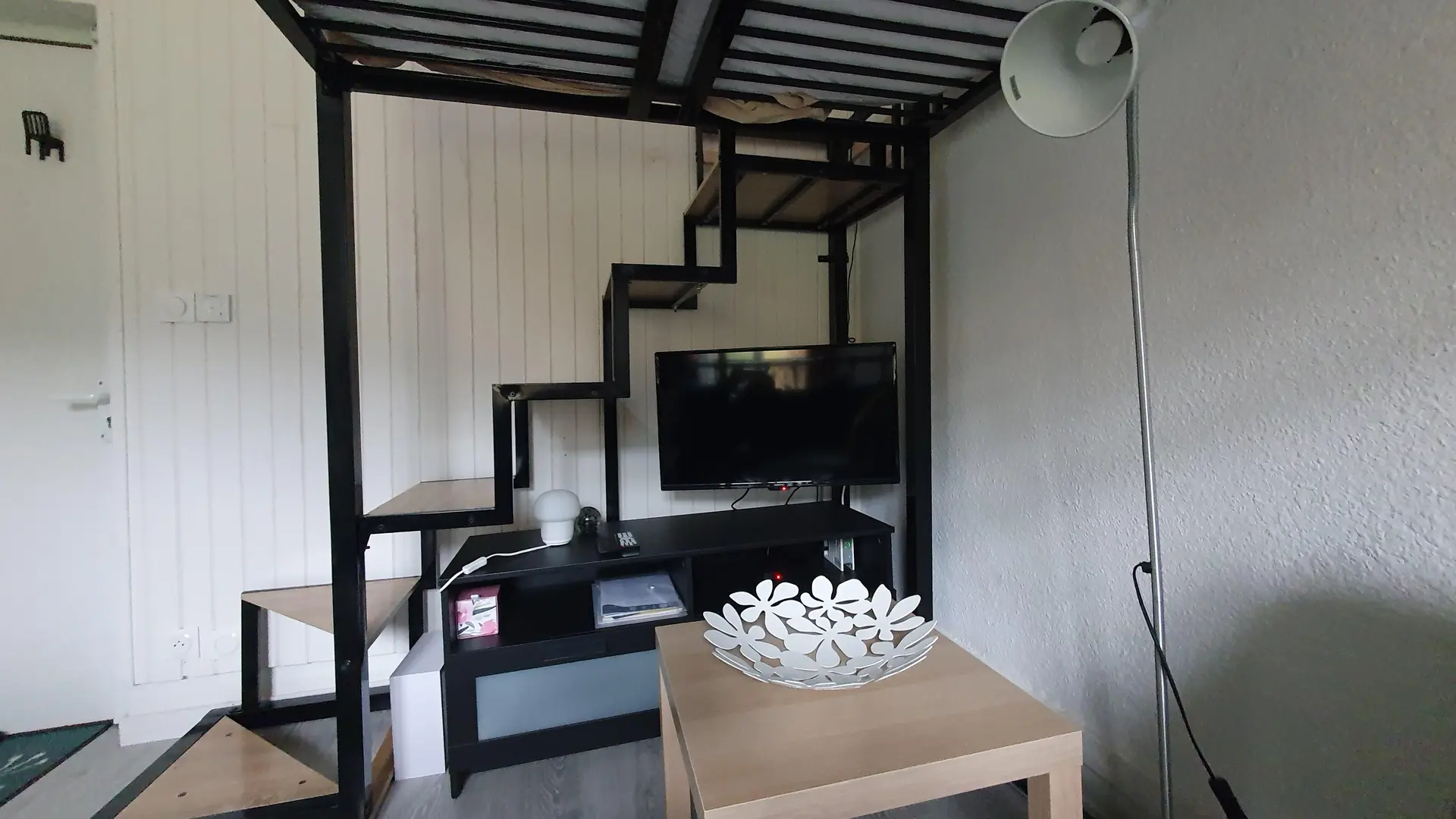 La pièce est un studio de style moderne caractérisé par une structure en métal noir supportant une mezzanine ou un lit superposé, avec un escalier visible, une télévision posée sur un meuble noir en dessous, et une table basse rectangulaire en bois clair au premier plan.