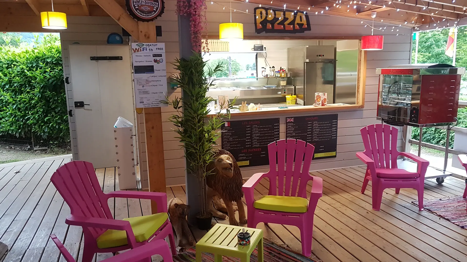 Un espace de restauration couvert en bois pour des pizzas à emporter, comprenant un comptoir et des tables avec chaises colorées, situé dans un environnement extérieur.