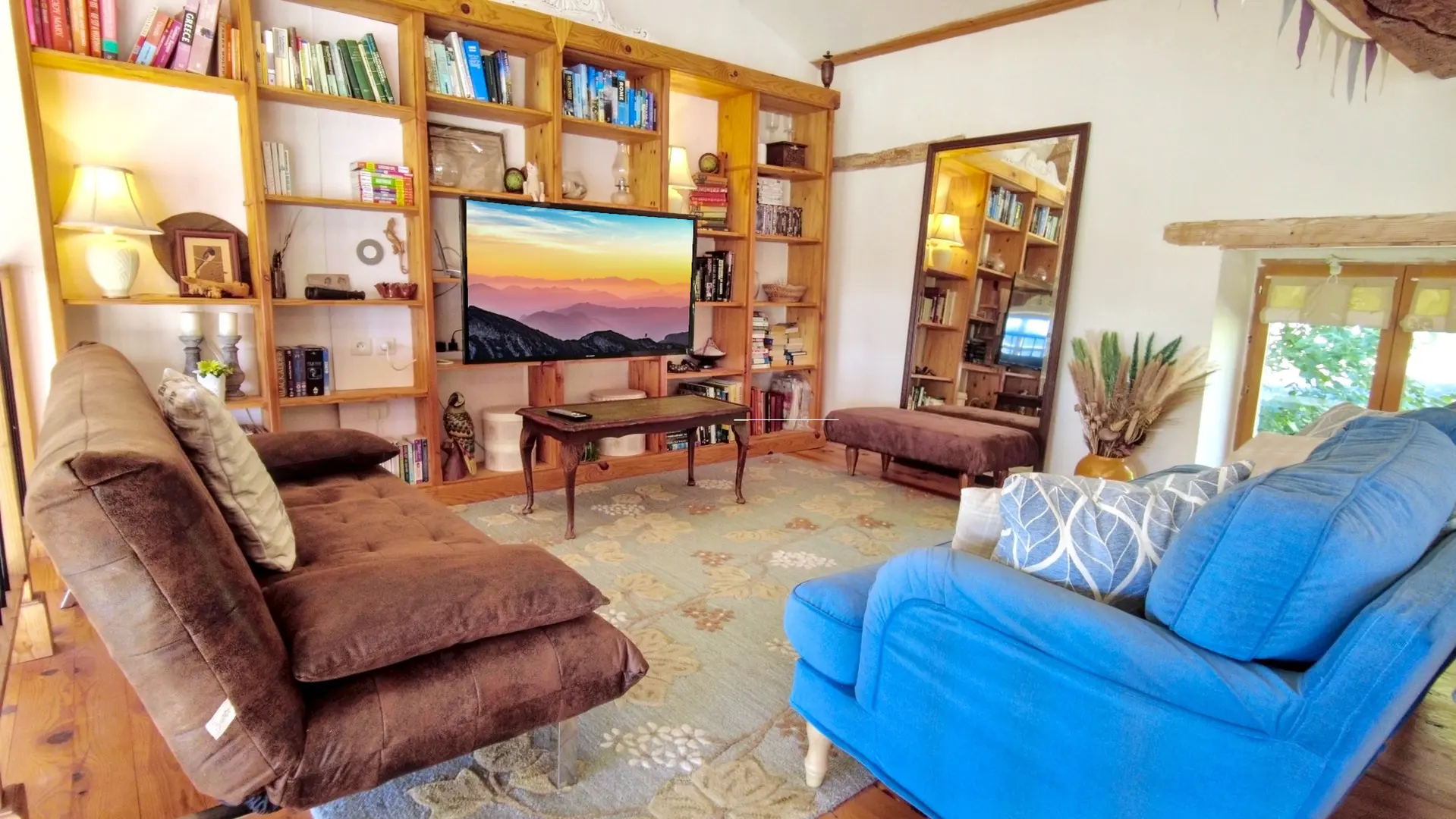 Un salon lumineux et accueillant, doté de planchers en bois. Deux canapés confortables sont orientés vers le centre : un canapé-lit marron foncé texturé sur la gauche, et un fauteuil ou un canapé bleu vif sur la droite, avec des coussins à motifs blancs et bleus. Entre les deux, un tapis à motifs floraux beiges et bleus couvre le sol. Au centre, une petite table basse en bois sombre de style antique est visible. Sur le mur du fond, de grandes étagères encastrées en bois clair remplies de livres et d'objets décoratifs entourent un écran de télévision plat qui affiche un paysage montagneux au lever ou au coucher du soleil. À droite, un grand miroir sur pied au cadre sombre reflète le reste de la pièce. La pièce présente des poutres en bois apparentes au plafond et une fenêtre à droite qui laisse entrer la lumière naturelle.