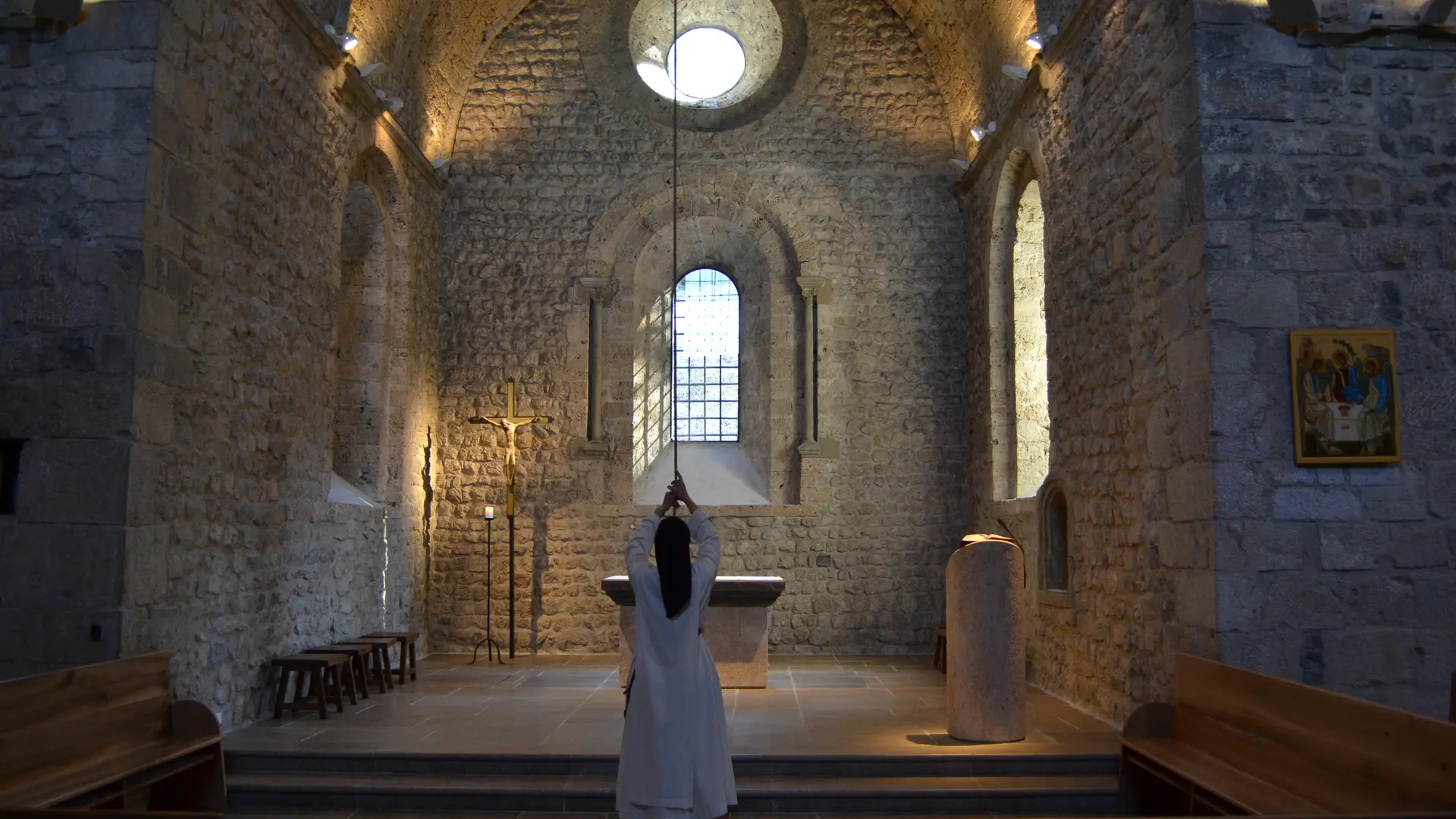 Au coeur de l'édifice éclairé d'une lumière filtrée par les vitraux, une personne en habit religieux s'apprête à tirer la ficelle reliée à la cloche de l'Église