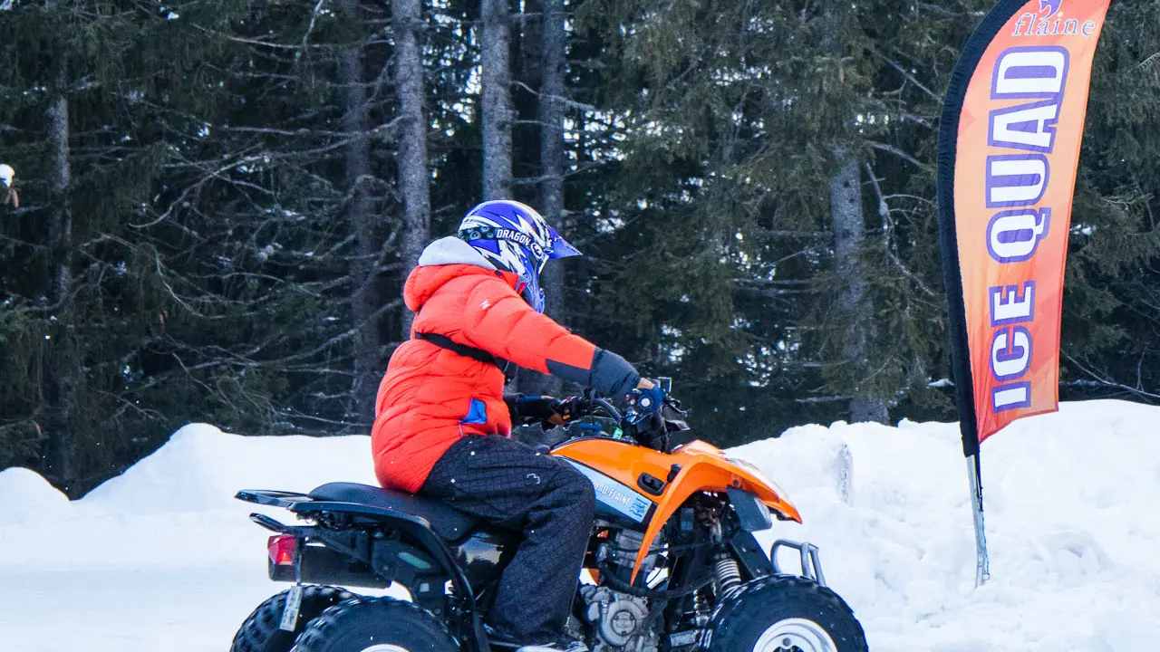 Une personne en veste orange et casque bleu conduit un quad noir et orange de profil. Le véhicule roule sur une piste enneigée devant une forêt de sapins.