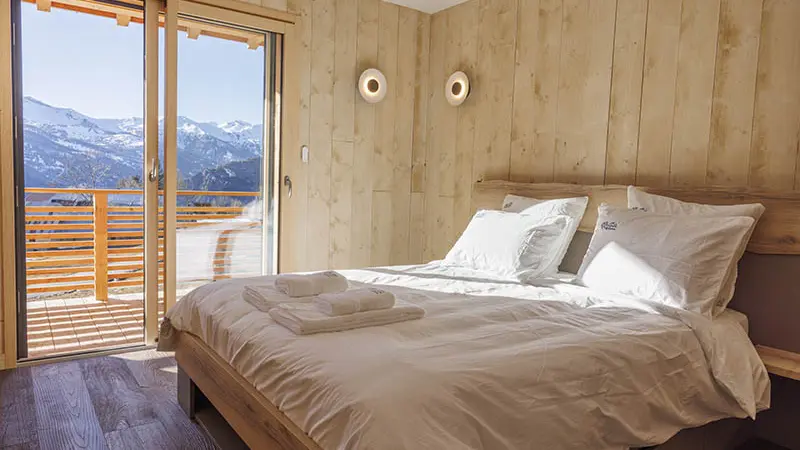 Chambre à coucher spacieuse et lumineuse avec lit double, ameublement bois et parquet, porte-fenêtre donnant sur un balcon en bois et un paysage enneigé