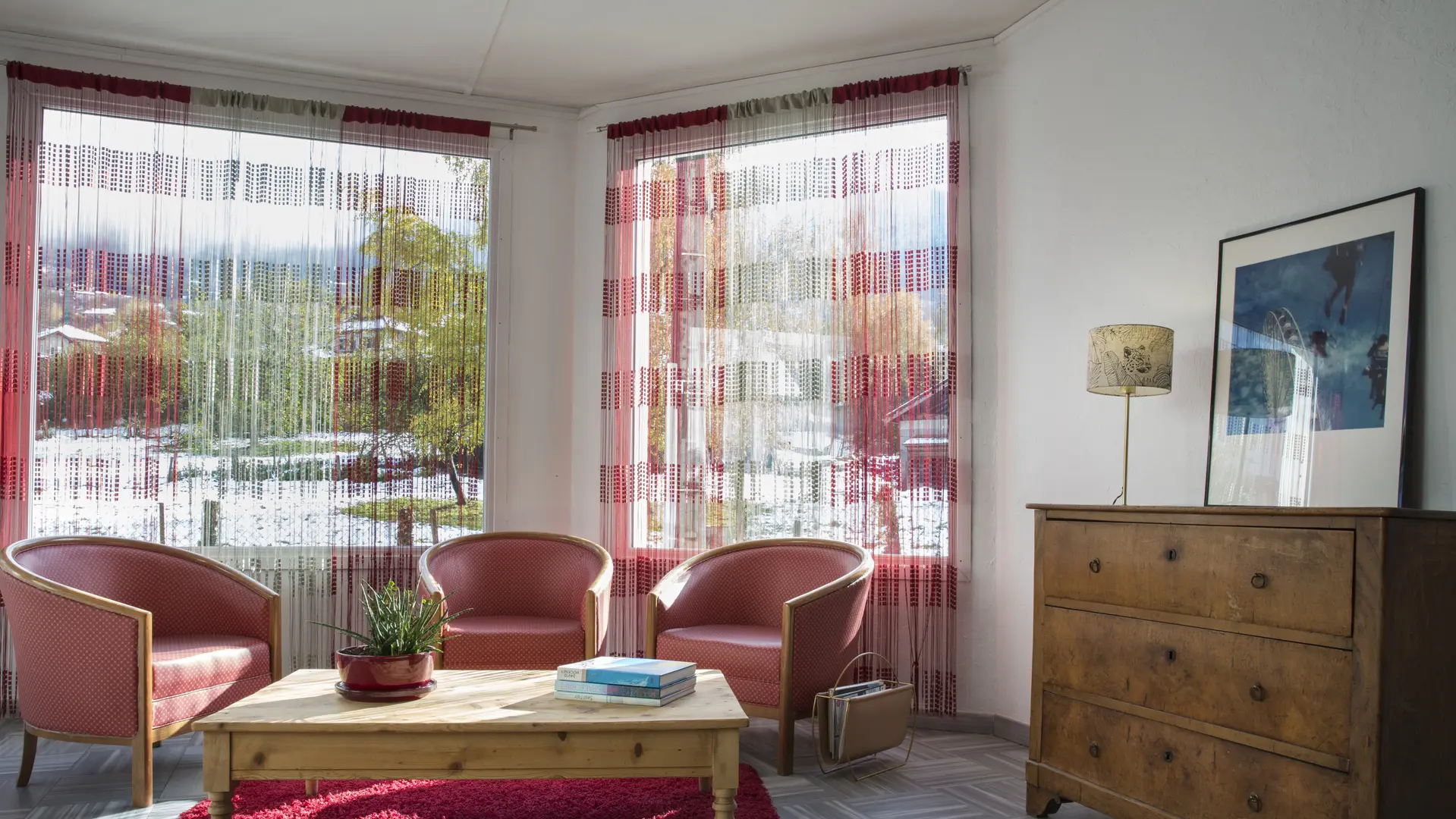 Le salon est équipé de plusieurs fauteuils rembourrés en tissu rouge, invitant à la détente et à la conversation. Une table basse en bois offre un espace pour poser des boissons ou des objets décoratifs.  La pièce bénéficie d'une belle luminosité grâce à de grandes fenêtres. Les rideaux légers filtrent la lumière naturelle et créent une atmosphère douce et apaisante.