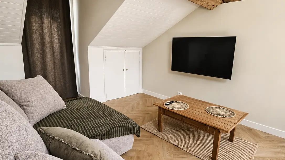 Un salon sous un plafond mansardé avec des poutres en bois apparentes, meublé d'un grand canapé d'angle en tissu clair, d'une table basse en bois posée sur un tapis et d'un écran de télévision accroché au mur.