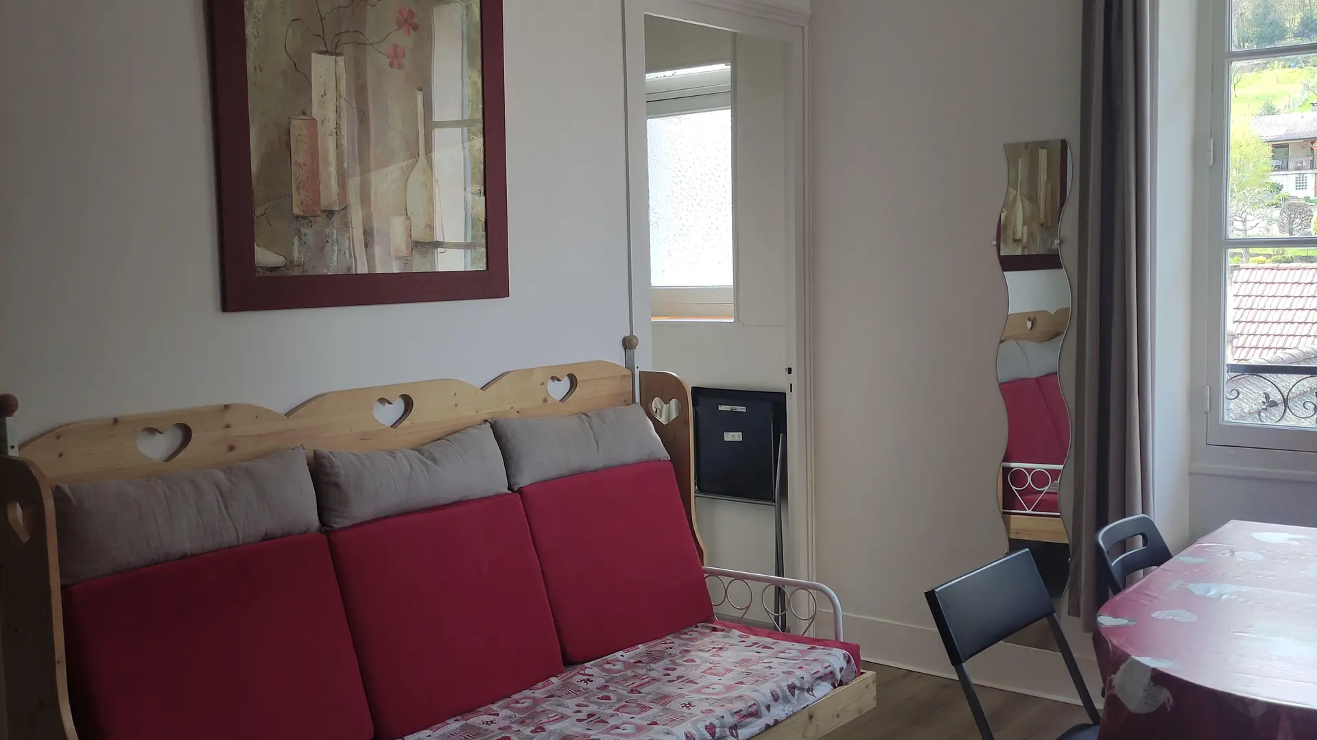 Un coin salon avec un canapé avec des coussins rouges et un coin repas avec une table ovale couverte d'une nappe rouge et des chaises, le tout dans une pièce avec un sol gris et une fenêtre sur la droite.