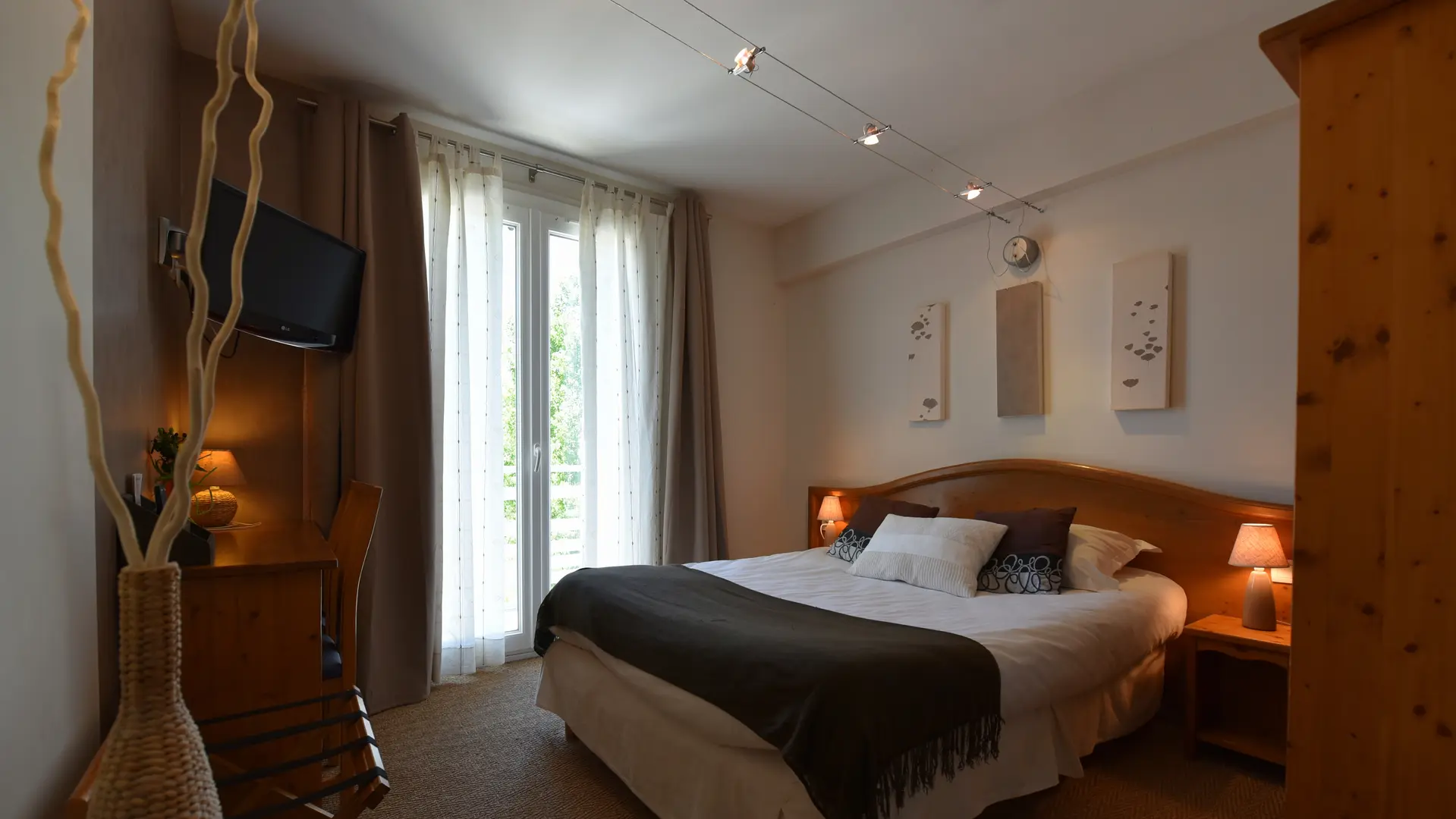 Chambre double chaleureuse équipée de deux lampes de chevet et d’une lampe de bureau allumées, de trois cadres décoratifs au-dessus du lit, d’une armoire, d’un porte bagage et d’un téléviseur. Trois spots orientables éclairés sur rail assurent un éclairage central. Vue sur la verdure par la porte-fenêtre