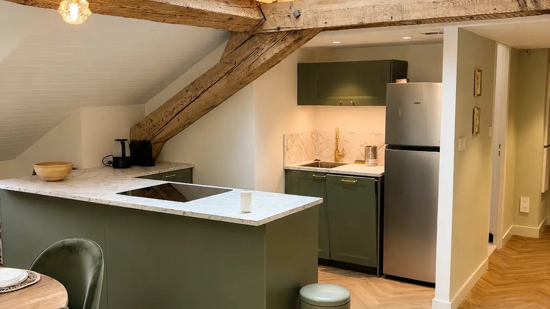 Une cuisine ouverte sous un plafond mansardé éclairé par une fenêtre de toit, équipée de meubles vert olive, d'un îlot central, d'un réfrigérateur en inox, de poutres en bois apparentes et d'un sol en parquet.