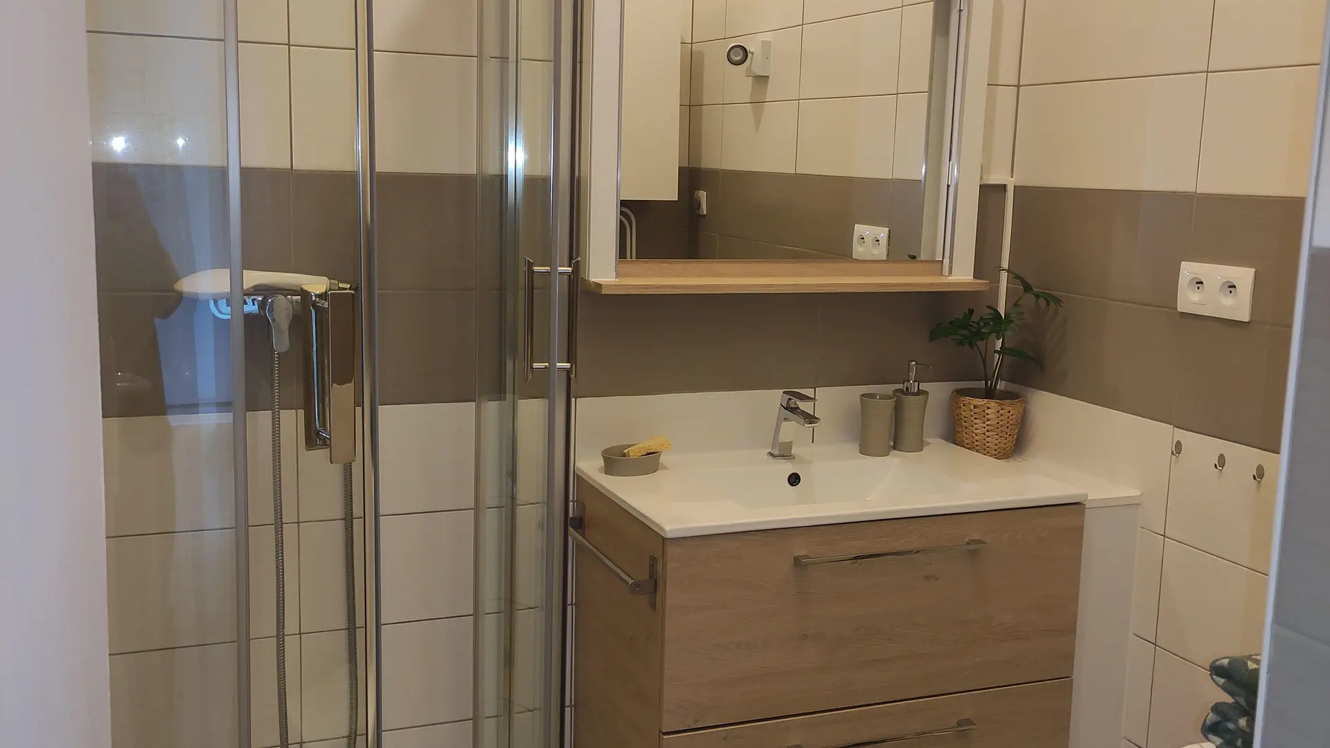 Une salle de bain avec une douche d'angle à paroi vitrée sur la gauche, un meuble-lavabo suspendu de couleur bois clair surmonté d'un miroir et d'une étagère, le tout dans une pièce aux murs carrelés beige clair et au sol foncé parsemé d'objets.